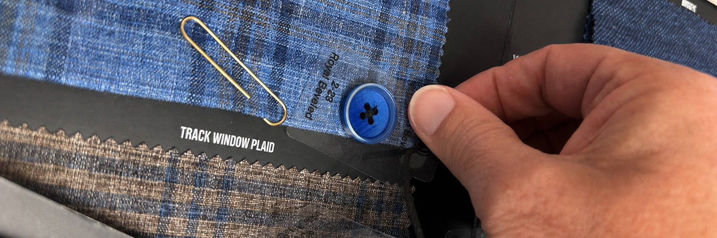 A close-up of a person's hand pointing at a blue plaid fabric swatch on a display card, with other fabric samples, buttons, and a paperclip visible.