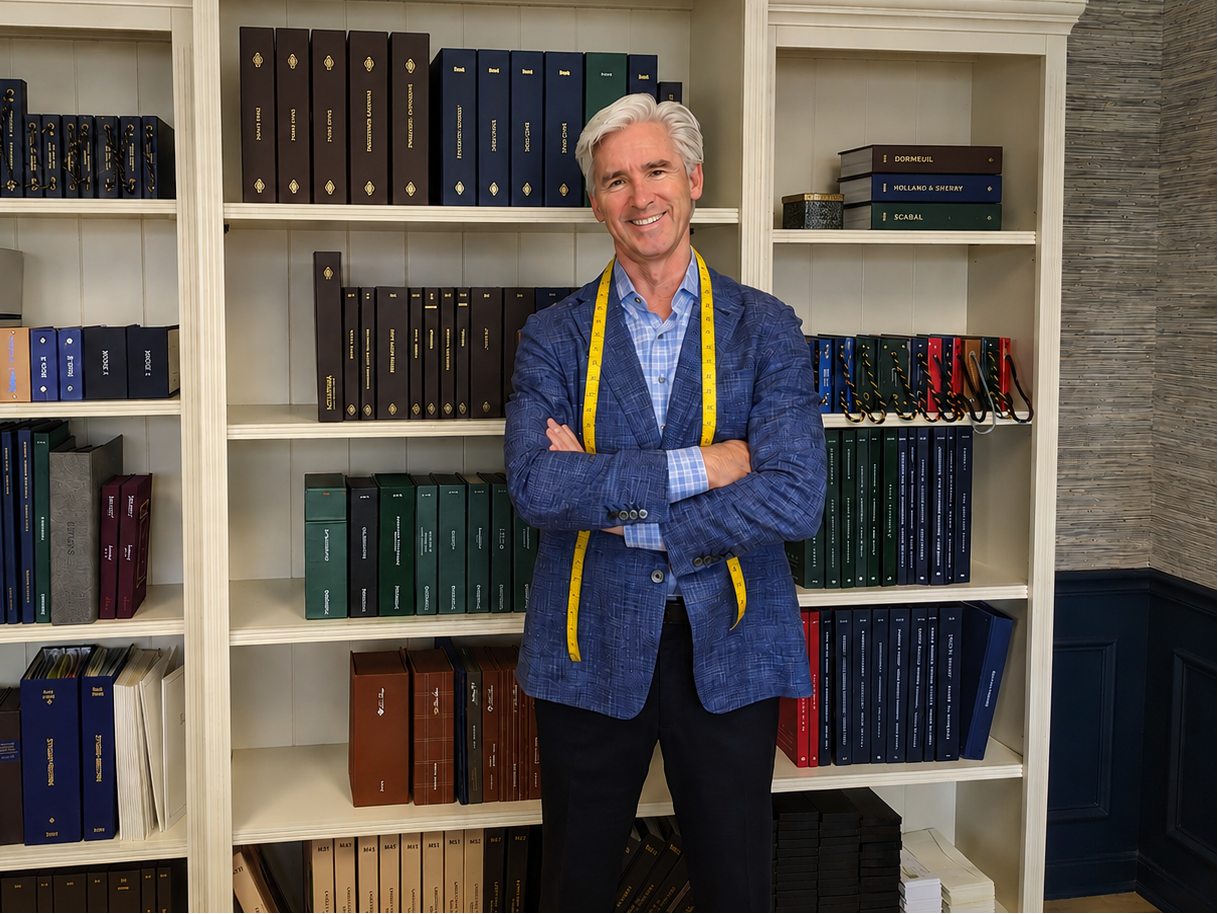 A smiling man with white hair stands in front of large cream-colored bookshelves filled with books. He is wearing a blue blazer with a yellow measuring tape draped around his shoulders and dark trousers.