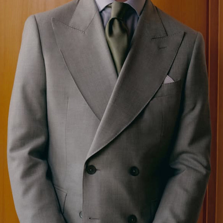 Light gray double-breasted suit crafted from Dormeuil luxury fabric, featuring broad peak lapels, an olive silk tie, and timeless bespoke-inspired tailoring.
