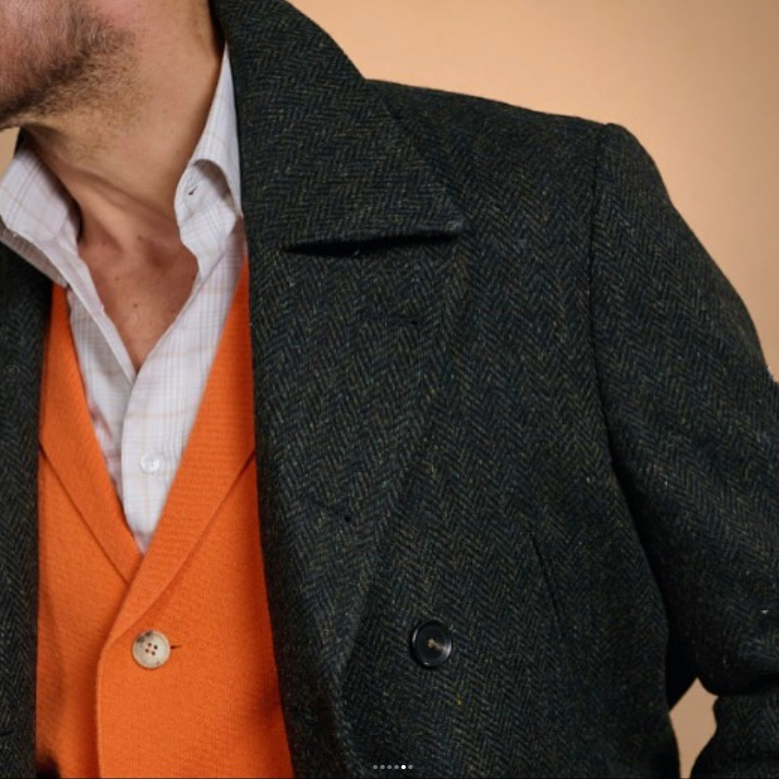 Detail of a Gladson luxury herringbone overcoat featuring premium wool fabric, classic tailoring, and layered styling with an orange cardigan for sophisticated seasonal menswear.