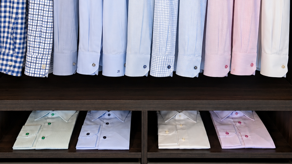 Dress shirts in blue, pink, and white solids, stripes, and checks, displayed with buttoned cuffs above folded shirts on dark wood shelves.
