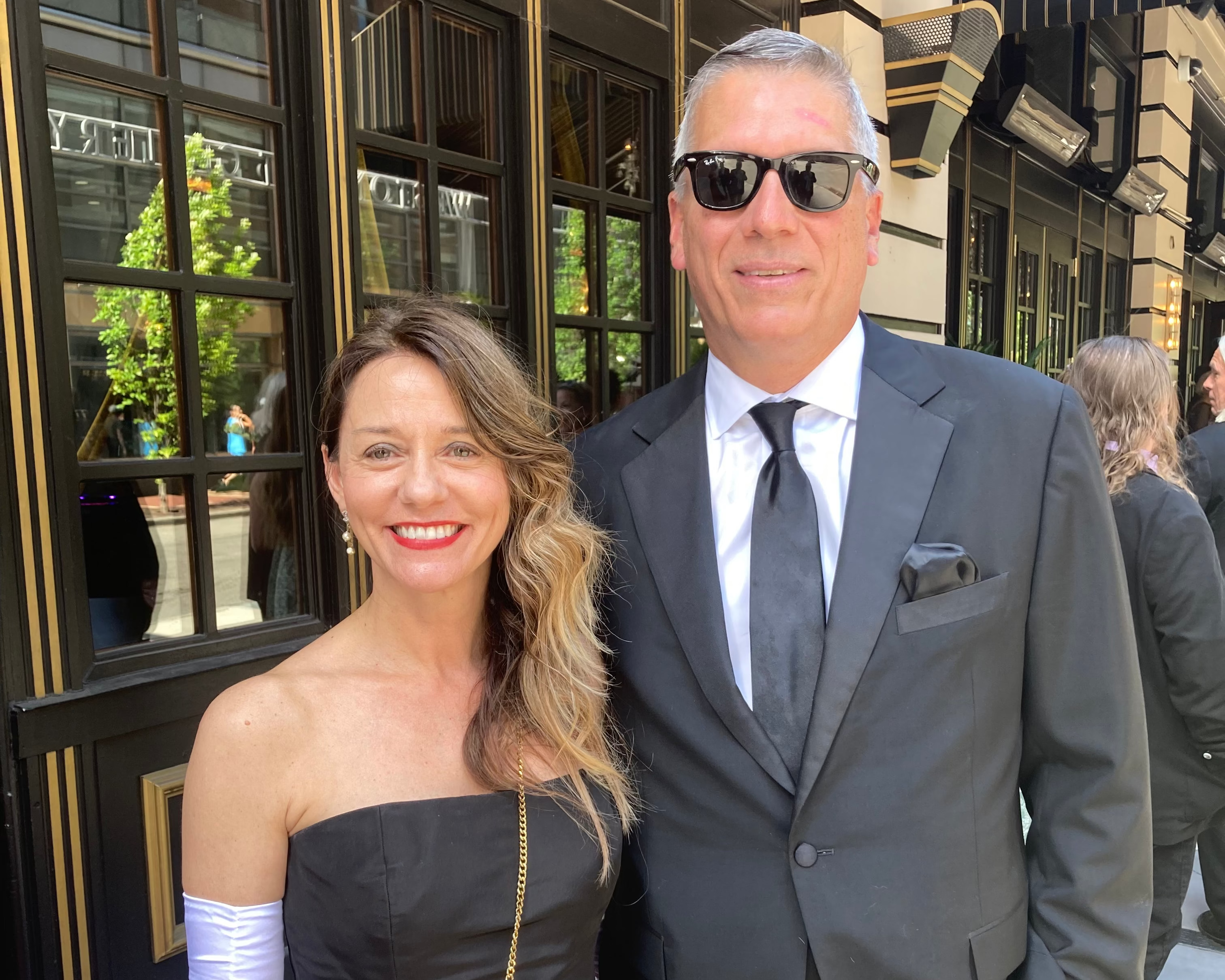 A man and a woman posing together outdoors in front of a luxury building. The woman is wearing a strapless black dress with long white gloves and a black crossbody bag with a gold chain strap.