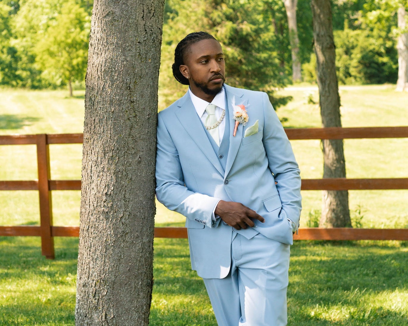 A man wearing a light blue three-piece suit, white dress shirt, and white bow tie leans against a large tree in a grassy park with a wooden fence in the background.